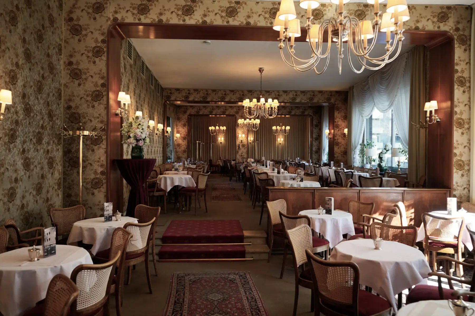 Kaffeehaus-Saal des Café Wahlen mit weiß eingedeckten Tischen, Kronleuchtern und gemusterter Wandbespannung